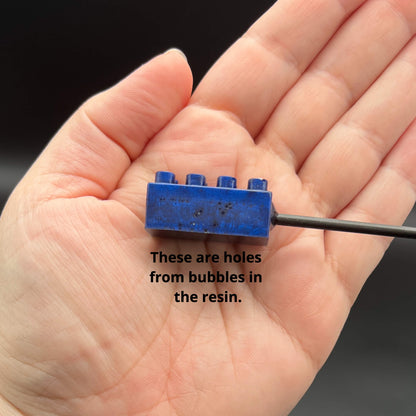 Close up side view of an imperfect blue building brick with tiny, shallow holes. The text reads, "These are holes from bubbles in the resin."