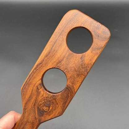 Close-up of a wooden paddle, Holy Terror, featuring two circular holes for added sensation.