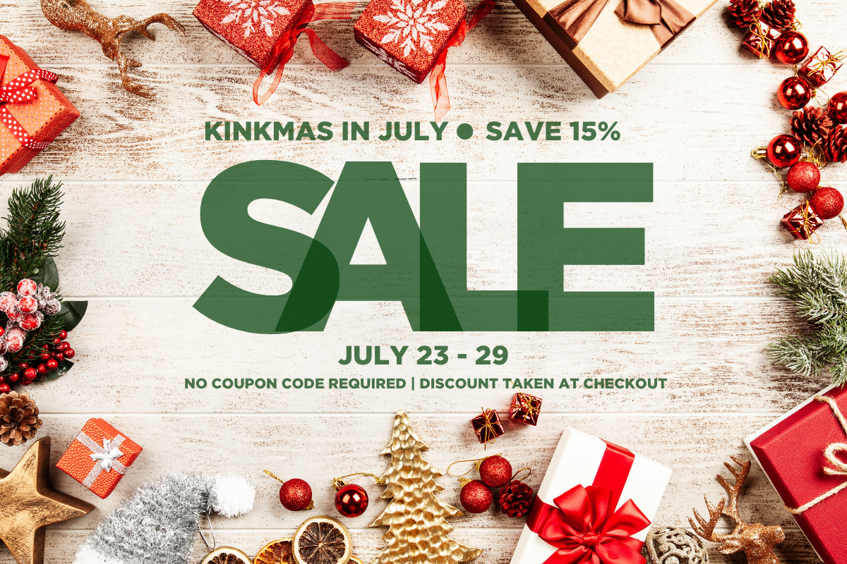 A promotional sale graphic for Kinkmas in July with large green text reading 'SALE' and dates 'JULY 23 - 29' on a whitewashed wood background, surrounded by Christmas decorations including wrapped gifts, red ornaments, pinecones, evergreen branches, and a small gold tree.
