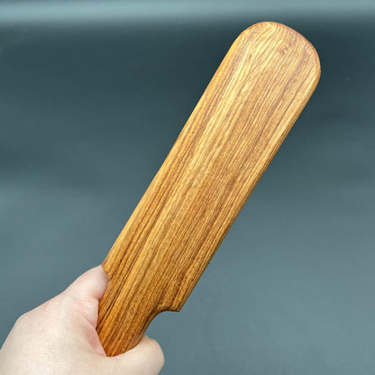 Hand holding a simple paddle made of natural wood, showcasing the Pounder with distinct grain pattern.