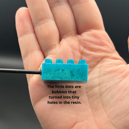 Close up side view of the teal building brick with small darker dots in the resin. The text reads, "The little dots are bubbles that turned into tiny holes in the resin."