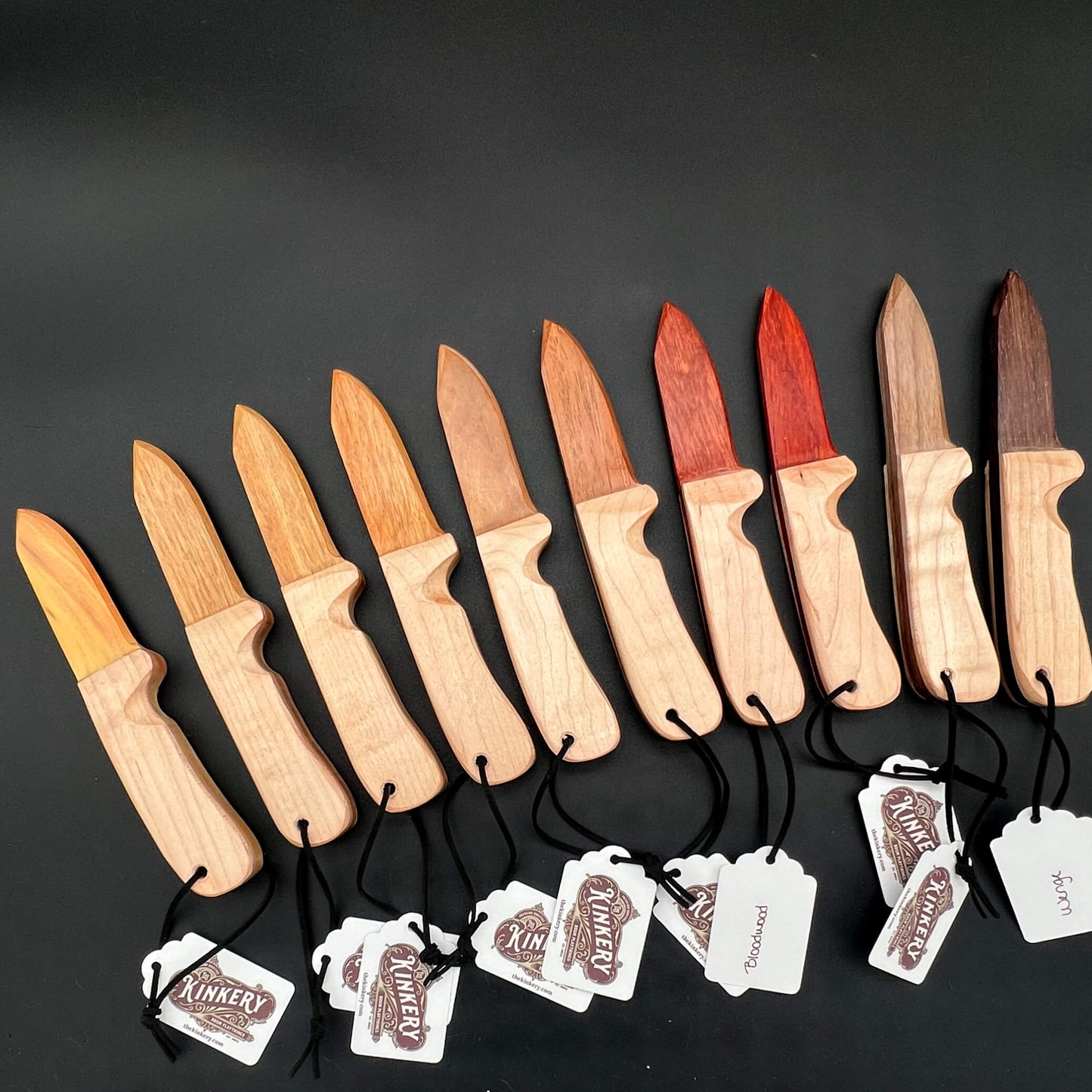 An array of wooden wax play knives with all different woods for the blades; the wooden knife is made in the style of an open switchblade knife.