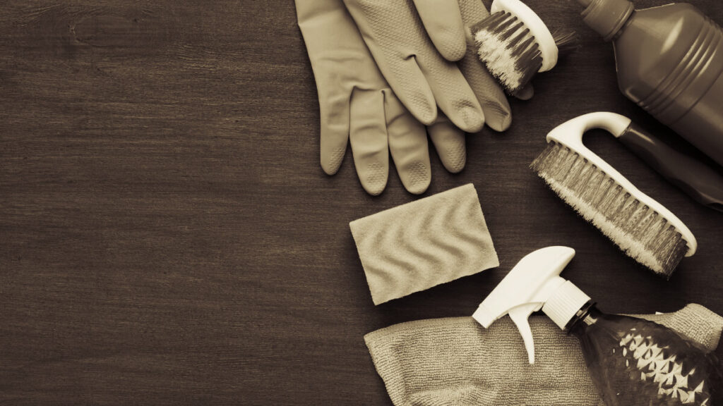 sepia toned image of cleaning supplies -- gloves, sponge, scrubber, spray bottle -- on wooden background for cleaning wooden kink toys