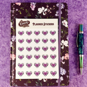 Sticker sheet of Loving BDSM logo planner stickers on top of floral purple planner next to turquoise pen on purple background