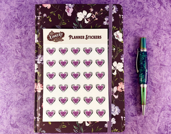 Sticker sheet of Loving BDSM logo planner stickers on top of floral purple planner next to turquoise pen on purple background