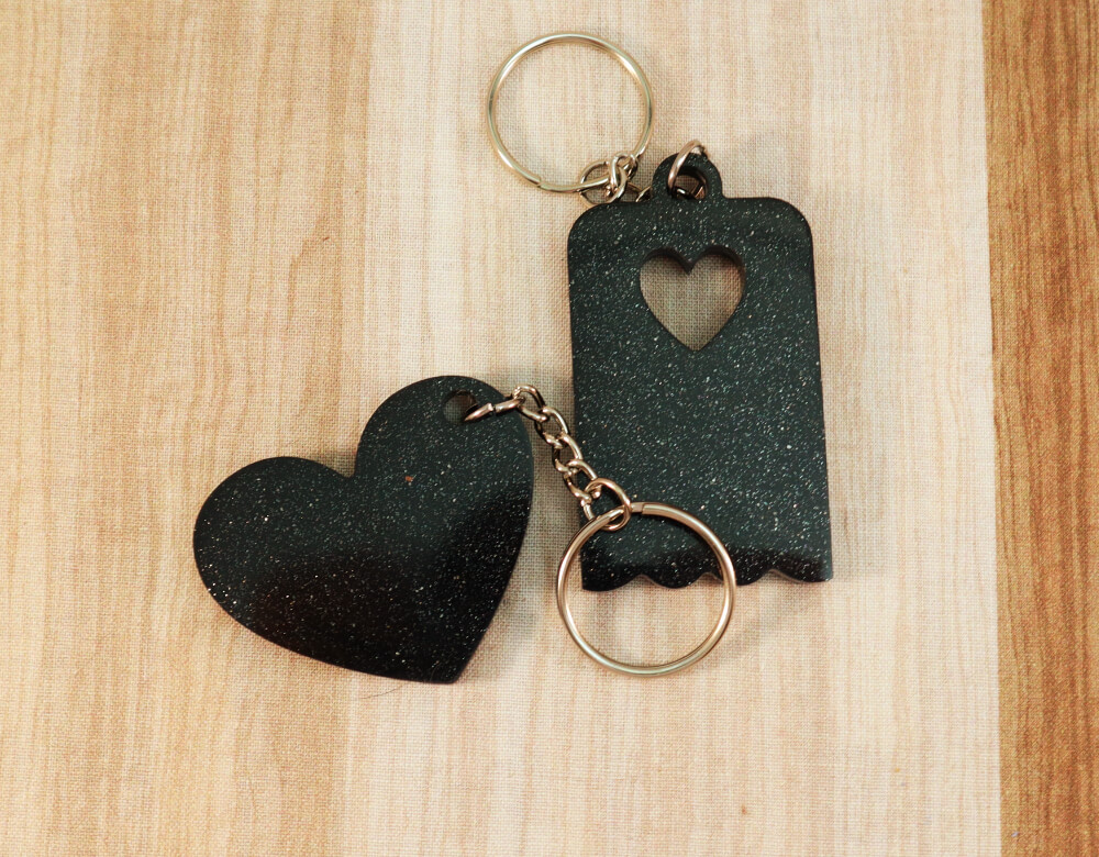 Two keychains in black resin with silver glitter; on left, heart, on right, scallop-edged rectangle with small heart cut-out