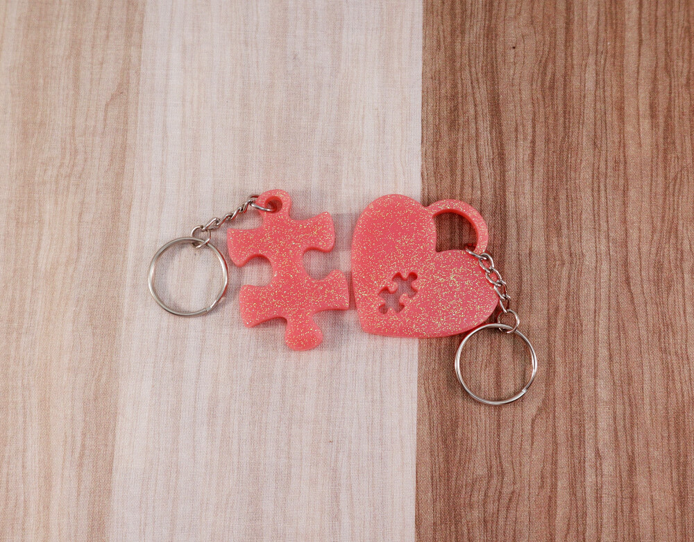 two keychain set in pink resin with multi-colored glitter; keychain on right is a puzzle piece; keychain on left is a heart lock with a puzzle piece cut-out