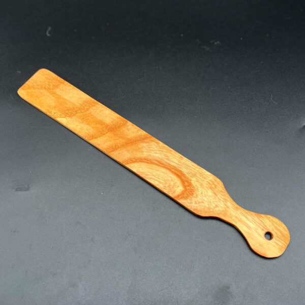 Narrow, thin, and long wooden paddle that looks like a ruler or a paint stir stick made in Ash, a yellow-brown wood with a strong curved grain pattern in a darker yellow-brown