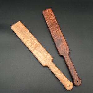 Two Ass Beater paddles - maple on the left, black walnut on the right. Maple is a light beige-brown wood. Black Walnut is a deep dark brown wood.