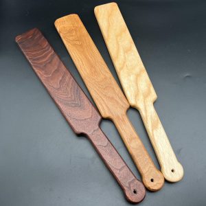 Three Ass Beater paddles in a variety of woods. Left to right: Black Walnut, Black Locust, and Ash. Each paddle is 18 inches long in total - 12 inch paddle length, 6 inch handle length.