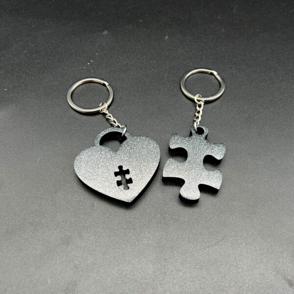 Two keychains made with black resin and silver glitter. On the left is a heart with a puzzle piece cut out at the bottom. On the left is a puzzle piece. Both have keychain hardware.