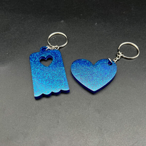 Two keychains made with blue resin and glitter. On the left is a rectangular piece with a scalloped bottom edge. On the right is a heart. Both have silver keychain hardware.