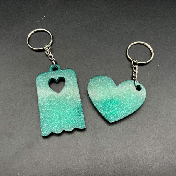 Two keychains made with green resin and glitter. On the left is a rectangular piece with a scalloped edge at the bottom and a heart cut out at the top. On the right is a heart. Both keychains have silver hardware.