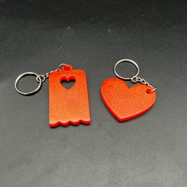 Two keychains made of orange resin and glitter. On the left is a rectangular piece with a heart cut out at the top and a scalloped edge on the bottom. On the right is a heart. Both have silver hardware.