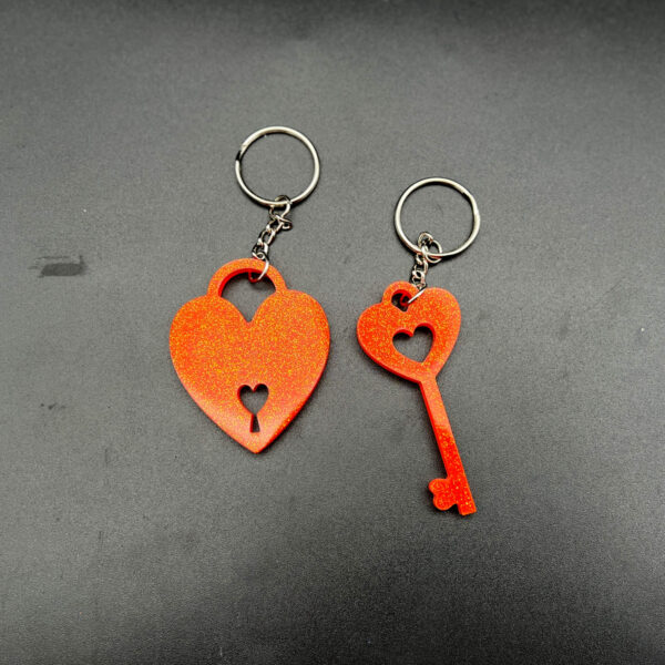 Two keychains made from orange resin and glitter. On the left is a heart with a lock cut out at the bottom. On the right is a key shape. Both keychains have silver hardware.