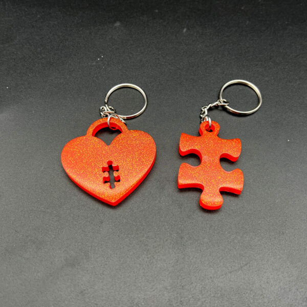 Two keychains made with orange resin and glitter. On the left is a heart with a puzzle piece shape cut out at the bottom. On the left is a puzzle piece. Both have silver hardware.