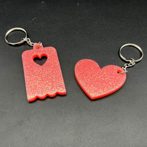 Two keychains made with pink resin and glitter. On the left is a rectangular keychain with a scalloped bottom and a heart cut out at the top. On the right is a heart. Both have silver keychain hardware.