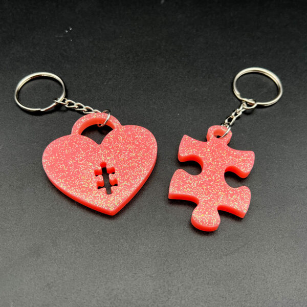 Two keychains made with pink resin and glitter. On the left is a heart-shape with a small puzzle piece cut out at the bottom. On the right is a puzzle piece. Both have silver hardware.