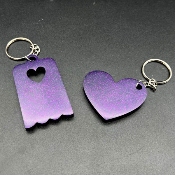 Two keychains made with purple resin and glitter. On the left is a rectangular keychain with a scalloped bottom and a heart cut out at the top. On the right is a heart. Both have silver keychain hardware.