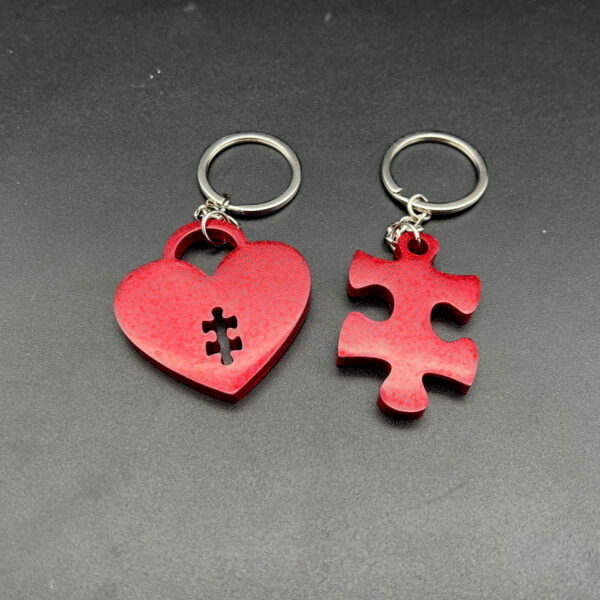 Two keychains made with red resin and glitter. On the left is a heart shape with a small puzzle piece shape cut out at the bottom. On the right is a puzzle piece. Both have silver keychain hardware.