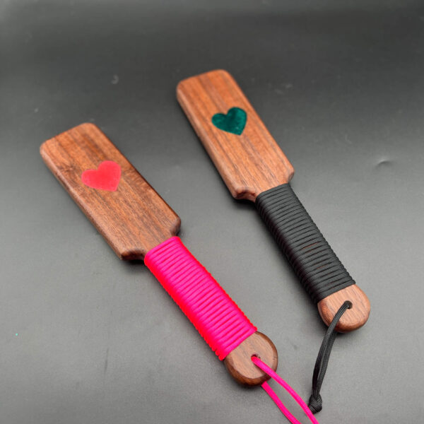 Two small resin inlay paddles in Black Walnut. On the left, a pink heart with pink paracord. On the right, a green heart and black paracord
