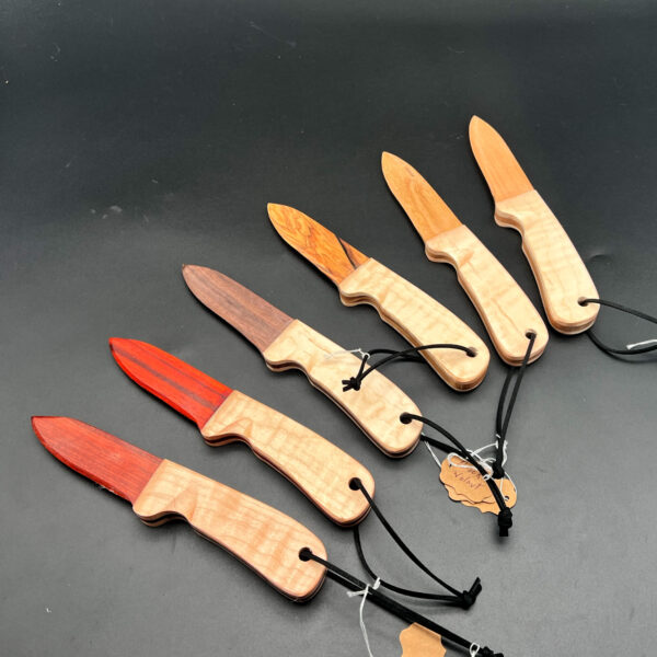 An array of wooden knives, all made with maple handles and a variety of blades. From left to right: bloodwood, paduak, black walnut, olive wood, cherry, and ash