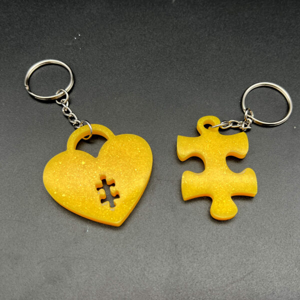 Two keychains made with yellow resin and glitter. On the left is a heart with a small puzzle piece shape cut out at the bottom. On the right is a puzzle piece. Both are made with silver keychain hardware.
