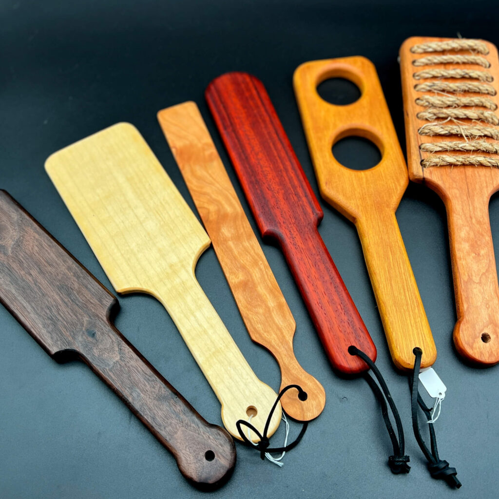 a variety of wooden spanking paddles made by the Kinkery. From left to right: Ass Beater Jr in black walnut; oversized hairbrush in maple; slapstick in cherry; Pounder in Paduak; Holy Terror in Osage Orange; and Rough Rider in cherry
