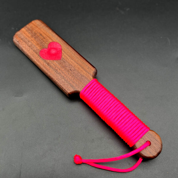 Small wooden paddle made of black walnut with pink resin heart in the center and pink paracord around the handle