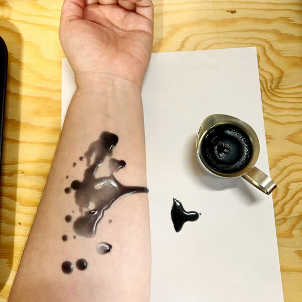 Black soy wax play candle on white paper next to Kayla's white arm. There are dried drops of black soy wax on her arm to show the color on skin -- this is an example of what it looks like on a light skin tone.