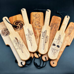 Variety of wood burned paddles showing multiple designs from swords to BDSM symbol and more