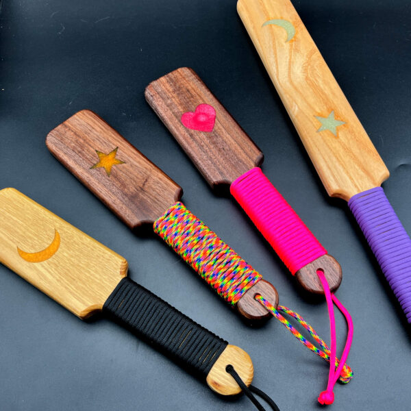 group of ready made resin inlay paddles. Left to right: Small black locust with yellow moon and black paracord; small black walnut with yellow star and rainbow paracord; small black walnut with pink heart and pink paracord; and large ash with glow in the dark moon and star with purple paracord