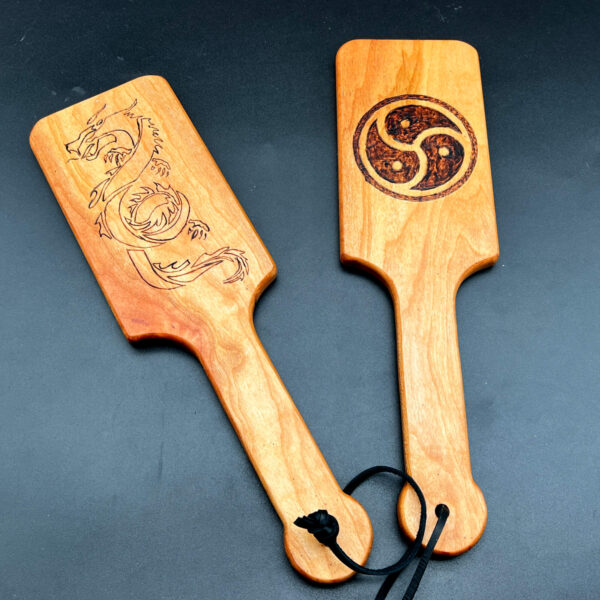 Two hairbrush style paddles with wood burned designs. Dragon on left, BDSM triskelion on right