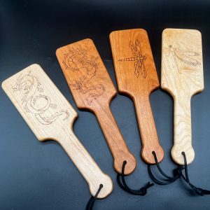 Four hairbrush style paddles with images wood burned into the center of the paddle. From left to right: maple paddle with a dragon, cherry paddle with a dragon, cherry paddle with a dragonfly, and maple paddle with a dragonfly.