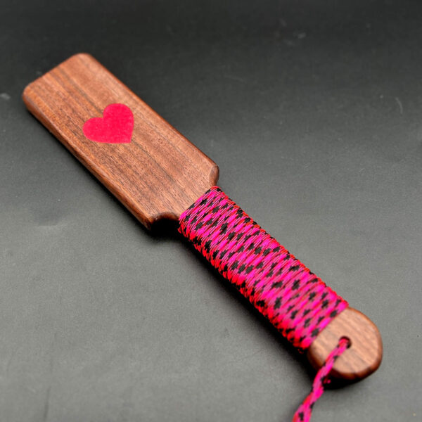 Small wooden paddle made of Black walnut with pink resin heart in the center and pink and black paracord around the handle