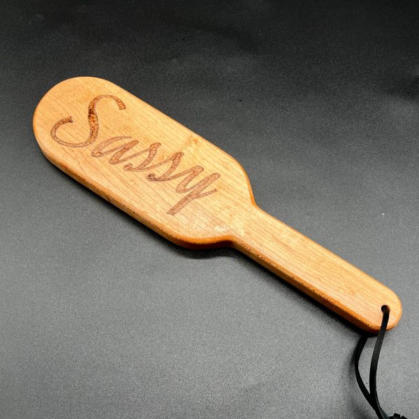 A 12 inch long wooden paddle with the word Sassy burned into the center of the paddle area. The paddle is 7 inches long and is in an oval shape. The handle is 5 inches long, 1.25 inches wide, and straight with no curves.