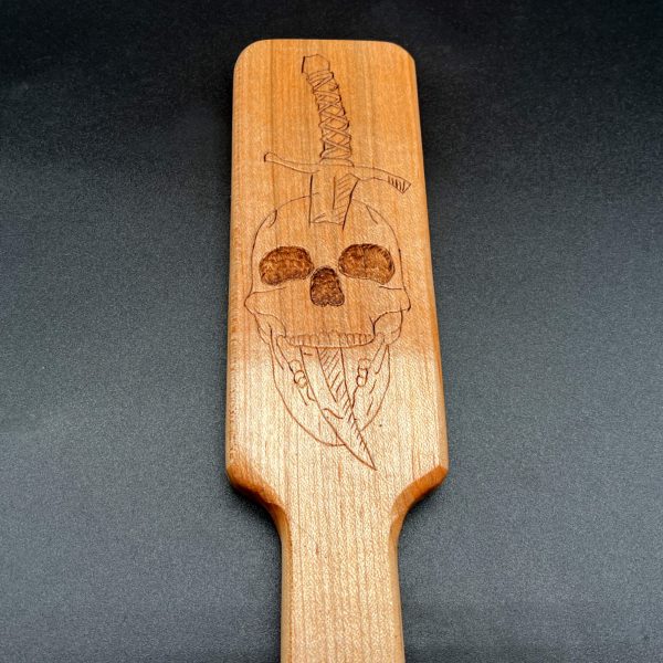 Close up view of a wood burned image in a wooden paddle: skull with dagger entering the top of the head and coming out through the mouth