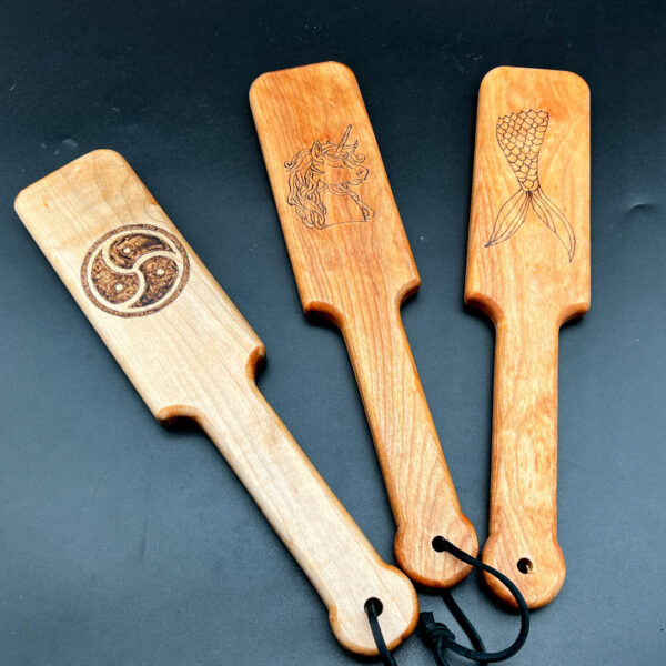 Three small wooden paddles with designs wood burned into center; left to right: BDSM triskelion, unicorn, mermaid tail