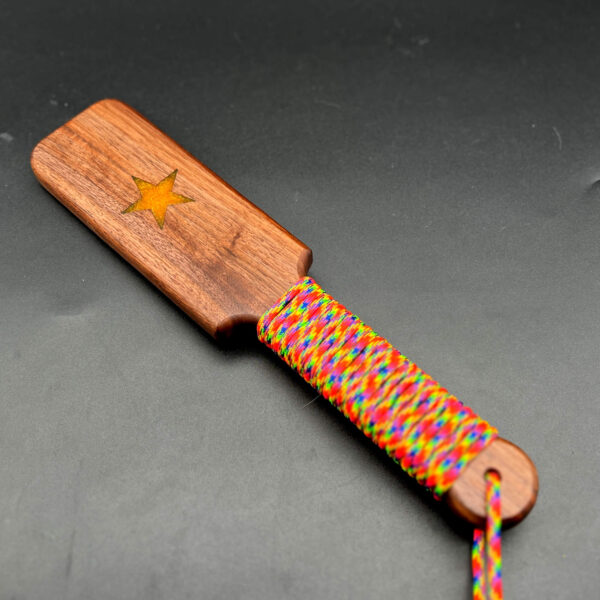 Small wooden paddle made of black walnut with yellow star in the center and rainbow paracord around the handle
