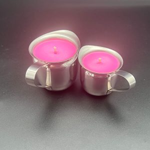 Two pink soy wax play candles side by side. The 5 ounce container is the on the left; the 3 ounce container is on the right. Both are made from stainless steel and have a handle and a pour spout.