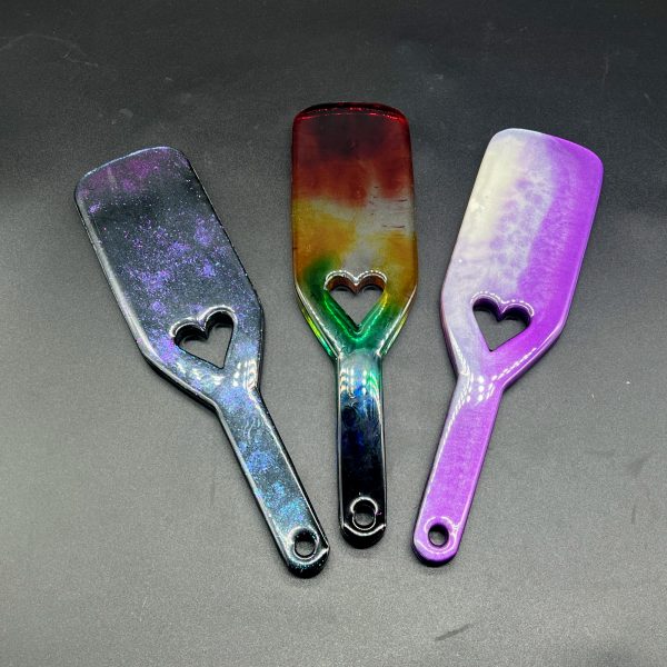 Three resin paddles side by side: purple to blue color shift in black resin, rainbow colors in clear resin, and purple and white resin combination